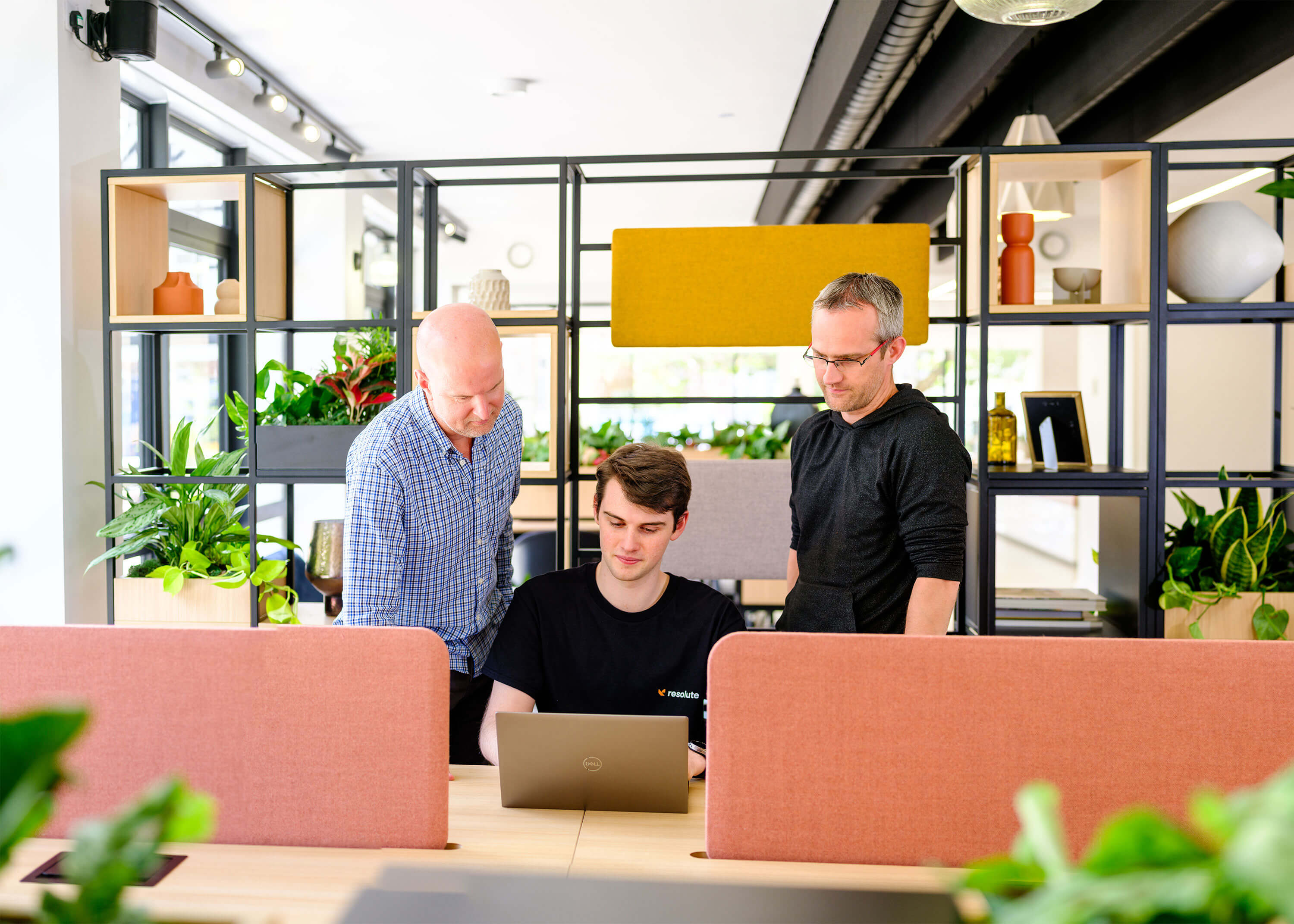Three Resolute Cyber team members collaborating around a laptop in a modern workspace with vibrant plants and decorative shelving.
